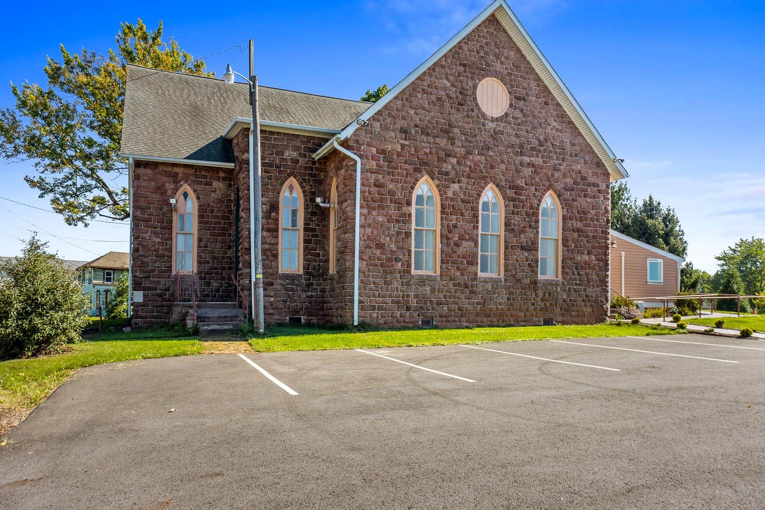 A stone building used as a wedding and event venue, with arched windows and a steep roof stands next to an empty paved parking lot on a sunny day.