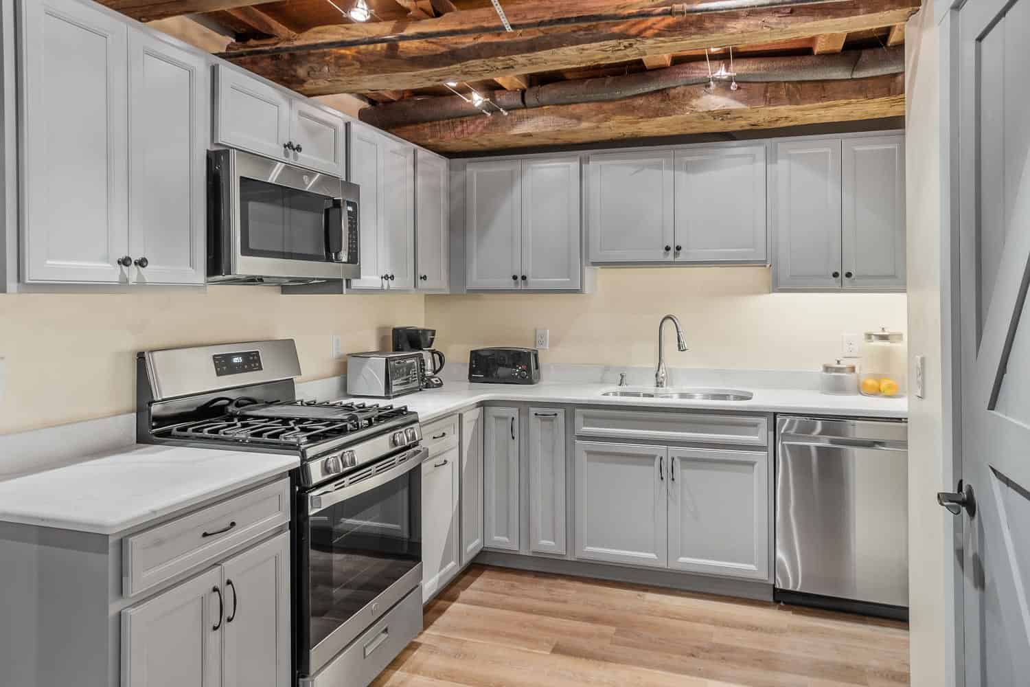 Venue-272-125 Modern kitchen with light gray cabinets, stainless steel appliances, exposed wooden ceiling beams, and light wood flooring.