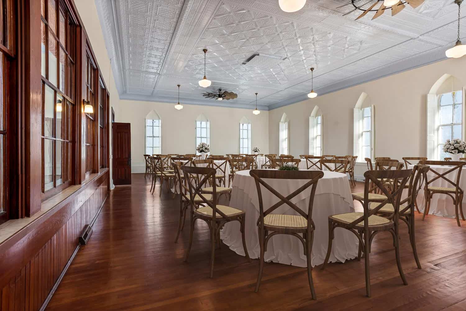 The Grace Room with wooden floors, several round tables covered with white cloths, wooden chairs, large windows, and a decorative ceiling.