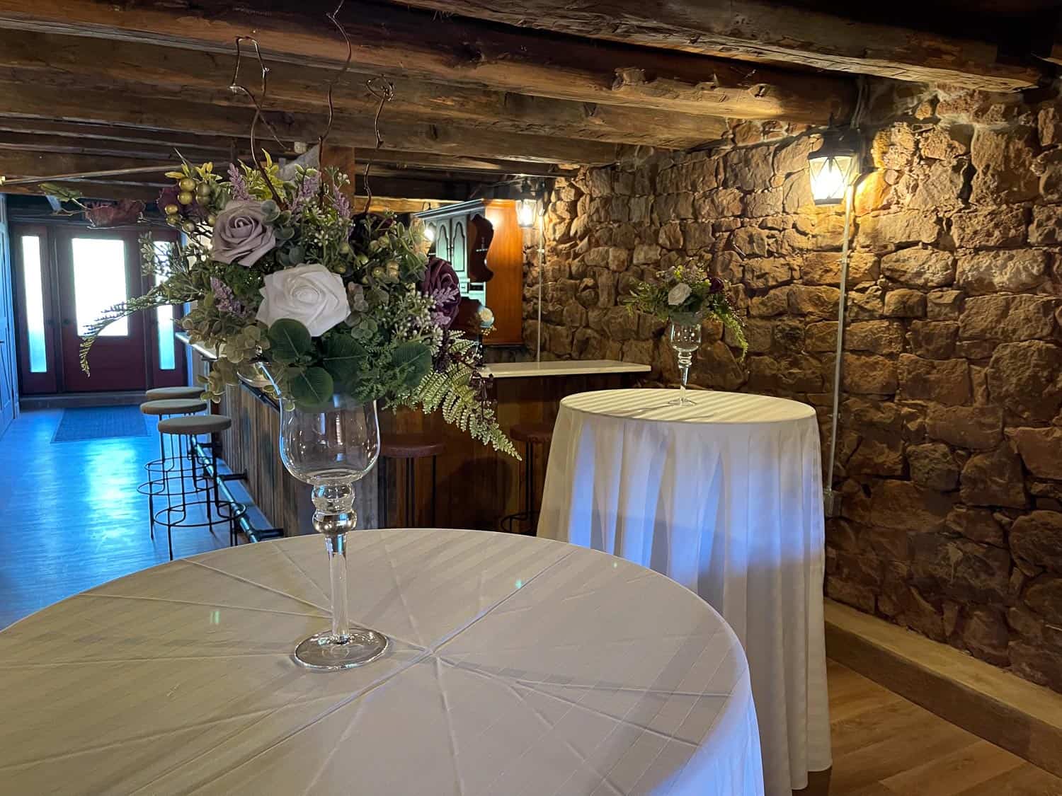Two cocktail tables with white tablecloths and floral centerpieces stand in the rustic Hearth Room, with stone walls, wooden beams, and soft lighting.